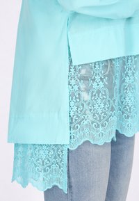 Una blusa de color turquesa claro con un dobladillo de encaje, que presenta intrincados patrones florales, superpuesta a unos pantalones vaqueros de mezclilla azul desgastada.