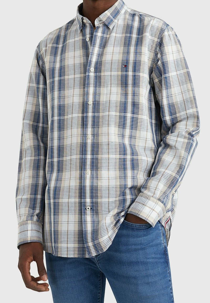 Camisa abotonada con un patrón de cuadros en azul y beige, hecha de tela liviana. Cuenta con cuello, mangas largas y un pequeño detalle de logo.