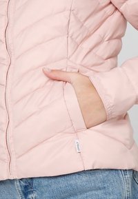 Jasnoróżowa pikowana kurtka z zamkiem błyskawicznym, kieszenią boczną i ściągaczami na mankietach. Charakteryzuje się ukośnym przeszyciem i małą metką z logo.