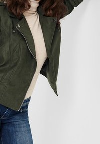 Veste en suédine faux vert olive avec une fermeture éclair à l'avant, des accents de boutons-pression et un design ajusté, superposée sur un col roulé beige.