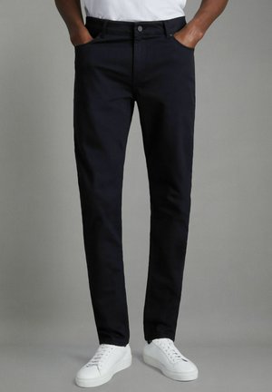 Pantalones de mezcla de algodón azul marino con piernas rectas, cinco bolsillos, cierre de botón y cremallera, y usados con zapatillas blancas.