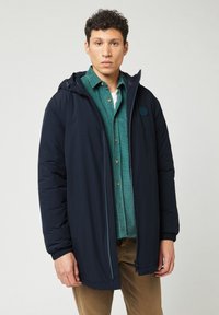 Chaqueta de padded en color marino con capucha desmontable, cremallera frontal, puños elásticos y una camisa verde texturizada debajo. Diseño casual adecuado para superponer.