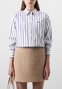 Witte shirt met paarse verticale strepen, met knopen aan de voorkant en een borstzak. Bruine vilt mini-rok, met een gladde textuur, met een hand die een beige tas vasthoudt.