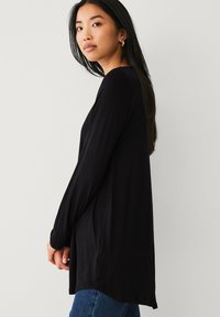 Haut noir à manches longues en tissu doux, doté d'une coupe décontractée, d'un ourlet courbé et de fentes latérales élégantes ; associé à un jean bleu.