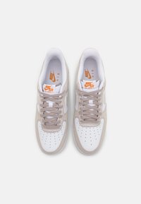 Nike Air Force 1 sneakers hebben een witte leren bovenkant met beige suède accenten, grijze veters, een geperforeerde neus en een oranje logo.