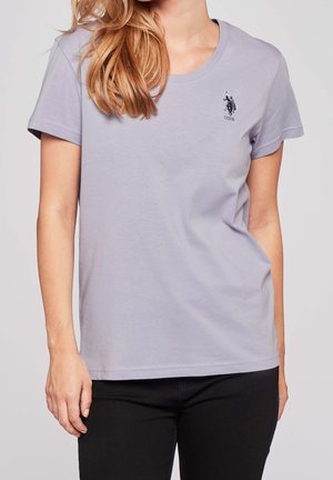 Vrouw draagt een lichtpaarse T-shirt met korte mouwen en een klein zwart USPA-logo op de linkerborst, gecombineerd met een zwarte broek.