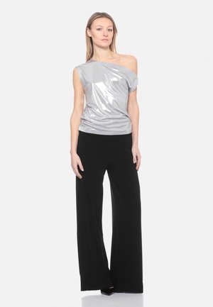 Donna in piedi con un top argento lucido monospalla e pantaloni neri a gamba larga su uno sfondo chiaro e semplice.