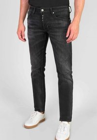 Hombre que lleva vaqueros negros ajustados con ligero desgaste y zapatillas blancas, de pie sobre un suelo gris claro.