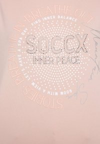 Maglietta rosa chiaro con design testuale circolare, dotato di motivi a punto e dettagli in argento. Il testo include "SOC CX", "INNER PEACE" e altre frasi.