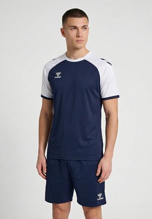 Mann trägt ein dunkelblau-weißes Hummel-Sportshirt und Shorts, steht mit entspannten Händen und schaut leicht zur Seite.