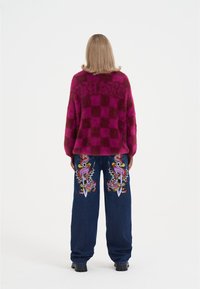 Plyšový růžový a bordó svetr s károvaným vzorem a nápisem "THANKS". Džíny s širokými nohavicemi v modré barvě s barevnými vyšívanými motivy ptáků a mečů.