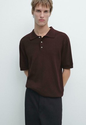 Giovane con capelli biondi corti che indossa una polo a maniche corte a coste di colore marrone scuro e pantaloni scuri, su uno sfondo bianco uniforme.