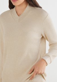 Beżowy sweter z prążkowanego dzianiny z dekoltem w serek, długimi rękawami i teksturowanym materiałem. Rękawy i dekolt w odcieniu nieco jaśniejszym beżu. Gładkie wykończenie.