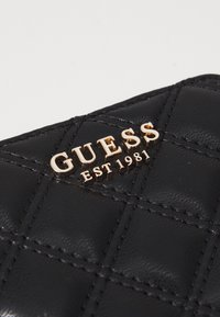 Svart kviltet lommebok laget av glanset skinn, med gull "GUESS EST 1981"-logo. Strukturert design med skrå sømmer.