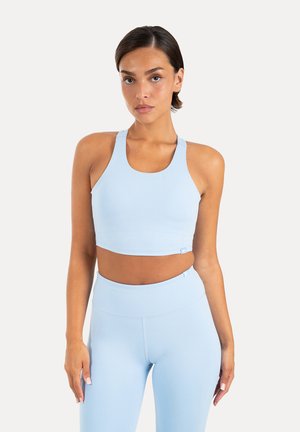 Junge Frau trägt ein hellblaues ärmelloses Crop-Top und hoch taillierte Leggings und steht vor einem schlichten weißen Hintergrund.