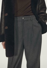 Grijs gestreepte broek met plooien, voorzien van een knoopsluiting. Gecombineerd met een donkere, gestructureerde jas met een zachte kraag.