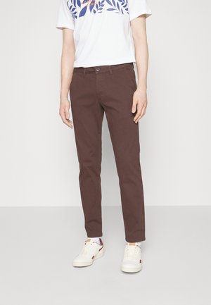 Personne portant un pantalon slim marron foncé, des baskets blanches avec des détails bordeaux, et une chemise blanche à manches courtes avec des imprimés de feuilles bleues.