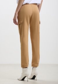 Pantaloni cargo beige con una vestibilità affusolata, polsini elasticizzati e due tasche laterali. Il materiale sembra morbido con una finitura leggermente testurizzata.
