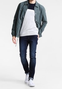 Leichte, dunkelgrüne Jacke über einem grau-blauen gestreiften Pullover, kombiniert mit dunklen Jeans und weißen Sneakers. Klassischer Freizeitlook.