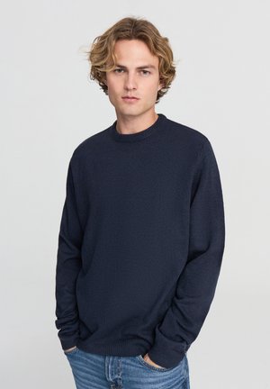 Maglione lavorato a maglia blu navy con scollatura tonda e maniche lunghe, caratterizzato da un tessuto texturizzato e una vestibilità morbida. Indossato con jeans azzurri.