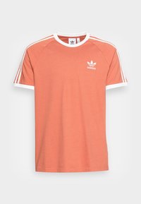 Meruňkový bavlněný tričko s bílým lemem, třemi bílými pruhy na ramenou a malým bílým logem Adidas na hrudi.