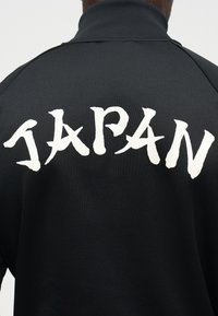 Sort jakke med hvide, stiliserede bogstaver, der staver "JAPAN" på tværs af den øvre ryg, båret af en person, der vender væk.