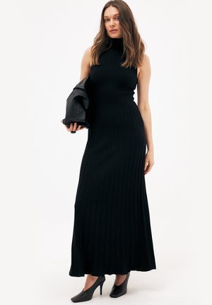 Vrouw draagt een mouwloze zwarte ribgebreide maxi-jurk, houdt een opgevouwen zwart jack in één hand vast en staat op zwarte pumps.