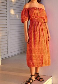 Robe orange à épaules dénudées avec une taille froncée, ornée de petits motifs géométriques. Portée avec des sandales noires à lanières.