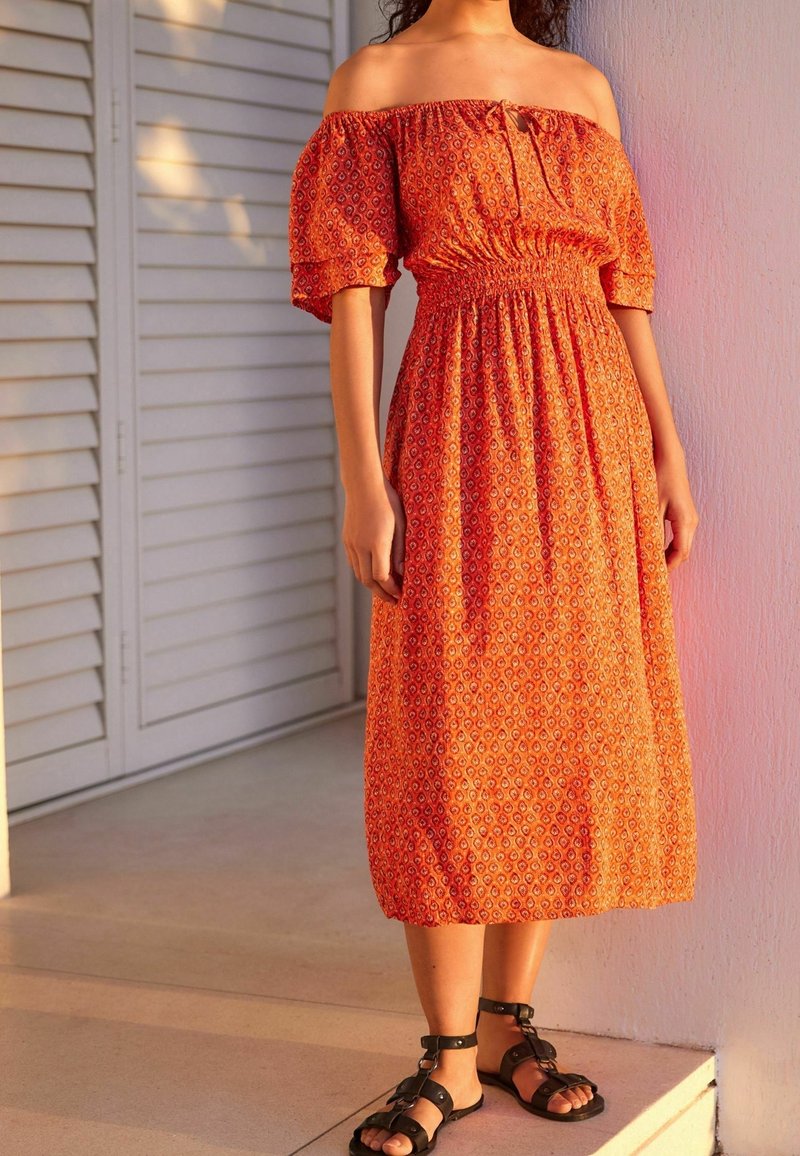 Robe orange à épaules dénudées avec une taille froncée, ornée de petits motifs géométriques. Portée avec des sandales noires à lanières.