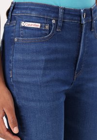 Jeans de mezclilla azul oscuro con un corte slim, que presentan una etiqueta de Calvin Klein en el bolsillo frontal y costuras decorativas en las costuras.