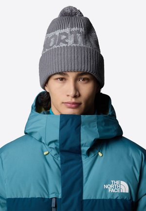 Grå strikket beanie med ribbet tekstur og en pom-pom på toppen; båret med en petrol- og marineblå hættetrøje med lynlås, der har et logo.