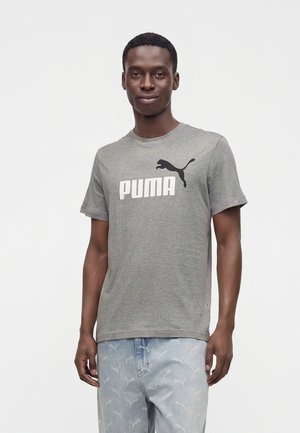 Νέος άνδρας που φοράει γκρι μπλουζάκι Puma και ανοιχτό μπλε τζιν με διακριτικό μοτίβο λογότυπου Puma, στέκεται μπροστά σε απλό λευκό φόντο.