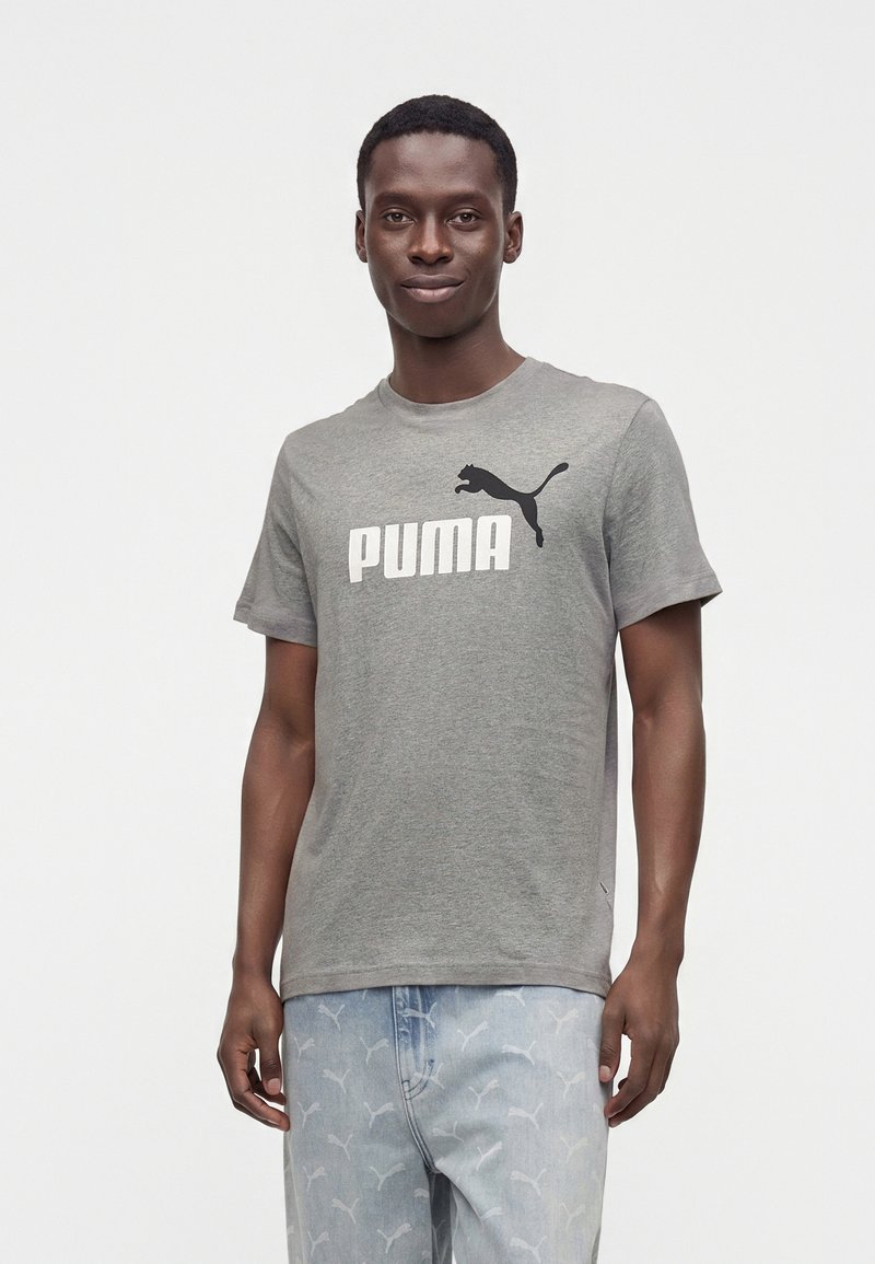 Νέος άνδρας που φοράει γκρι μπλουζάκι Puma και ανοιχτό μπλε τζιν με διακριτικό μοτίβο λογότυπου Puma, στέκεται μπροστά σε απλό λευκό φόντο.