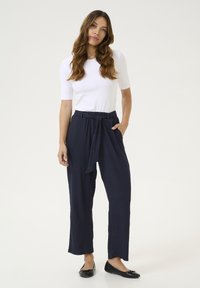 Bílý, těsný top v kombinaci s volnými tmavomodrými kalhotami s vázacím páskem a bočními kapsami. Černé ploché boty dokončují outfit.
