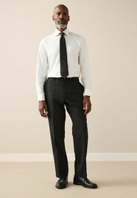 Fehér ing, mintás fekete nyakkendő, fekete nadrág és fekete cipő. Semleges háttér sima textúrával. Formális öltözködés.