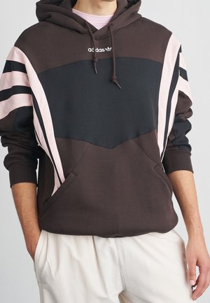 Personne portant un sweat à capuche marron avec des rayures diagonales noires et roses sur les manches et la poitrine, associée à un pantalon blanc, mains dans les poches.