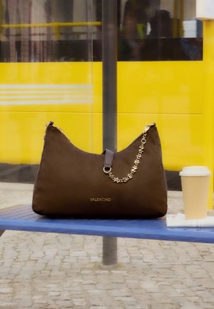 Кафява дамска чанта Valentino с детайли от златна верижка на синя пейка, кафе за вкъщи, жълт автобус размазан на заден план.