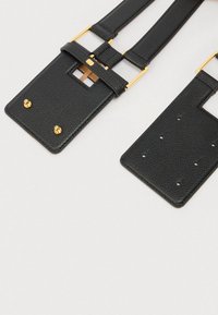 Μαύρη δερμάτινη ζώνη με φαρδιά ορθογώνια άκρα, καρφιά σε χρυσό τόνο και τετράγωνα ανοίγματα σε ανοιχτό γκρι φόντο.