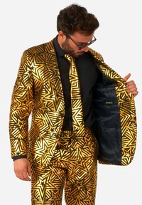 Zelta rakstainā uzvalkā ir zvaigžņu un ģeometrisko dizainu elementi ar melniem akcentiem. Iekšpuse ir izklāta ar tumši zilu audumu, kurā redzama zīmola etiķete.