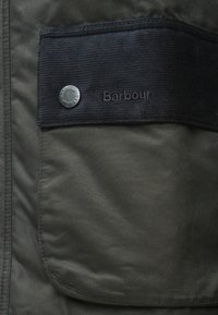 Sötétzöld kabát mankós zsebbel. Ezüst gombbal, amelyen a "Barbour" logó és dombornyomott felirat található a zseben. Texturált anyag.