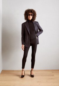 Blazer noir à rayures sur un col roulé noir, associé à des leggings noirs et des chaussures à talons. Le modèle a des cheveux bouclés et des lunettes de soleil.