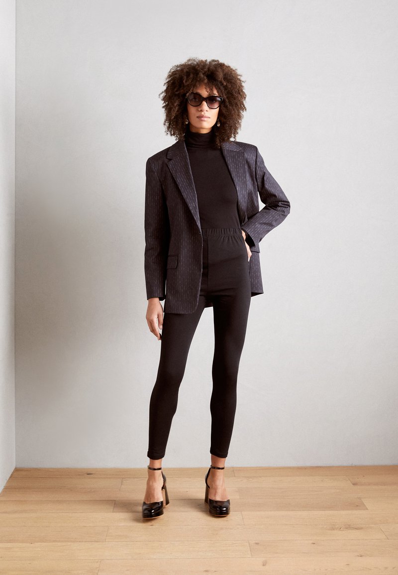 Blazer noir à rayures sur un col roulé noir, associé à des leggings noirs et des chaussures à talons. Le modèle a des cheveux bouclés et des lunettes de soleil.