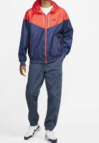 Nike windbreaker with red and dark navy color blocks, featuring a hood, zippered pockets, elastic cuffs, and matching navy cargo pants.