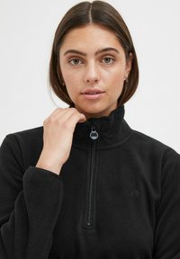 Pull en polaire noire avec col montant zippé, arborant un logo sur la poitrine et des poignets côtelés. Texture douce, tissu léger.
