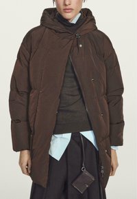 Brown puffer coat with a hood and front button closure, featuring a quilted design. Worn over a dark sweater and light blue shirt.