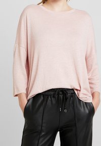 Jasnoróżowy, oversize sweter z miękkiej dzianiny, z opadającymi ramionami i luźnym krojem, zestawiony z czarnymi, skórzanymi spodniami z troczkami.