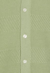 Jasnozielony sweter z dzianiny z teksturowanymi pionowymi i ukośnymi paskami, z centralnym zapięciem na guziki i okrągłymi, białymi guzikami.