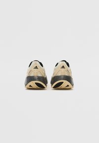 Des chaussures de sport beiges avec un dessus en maille texturée, des accents noirs et une semelle robuste et profilée avec une semelle grise et un logo.