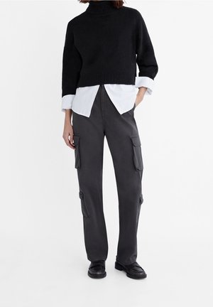 Osoba v černém zkráceném roláku přes bílou košili s ohrnutými rukávy, tmavých cargo kalhotách a černých mokasínech.