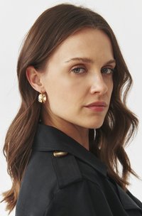 Gold hoop earrings with an abstract design, featuring a polished surface and a shiny texture, worn by a person with medium-length hair.